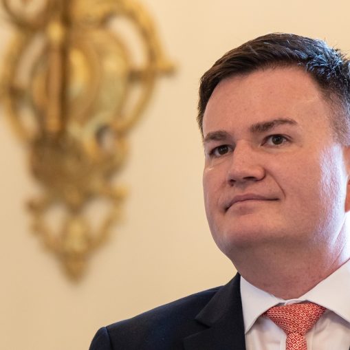 Arti kadro ir kairėje pusėje matyti dalis vyro veido ir krūtinės, dėvinčio tamsų švarką ir baltus marškinius su raudonais raštais.