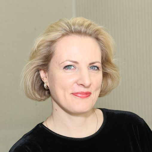 Šviesiaplaukė moteris su mėlynais akių kontūrais ir raudonais lūpų dažais dėvi juodą palaidinę.