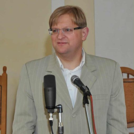 Vyras su akiniais ir šviesiu kostiumu kalba į mikrofoną.