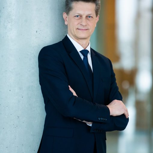 Vyras tamsiai mėlynu kostiumu su mėlyna peteliške stovi, atsiremijęs į pilkos betoninės kolonos ir yra sudėjęs rankas ant krūtinės.