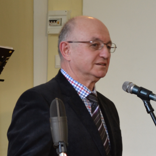 Baltaodžiai akiniai ir šviesiaplaukiai vyras, dėvintis švarką ir languotus marškinius, kalba į mikrofoną.