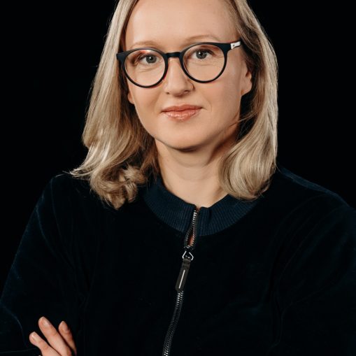 Moteris su akiniais ir plaukais kaip saulės spinduliai, pasitempusi ir pasitikinti, juodame fone.