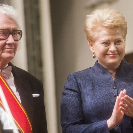 Vyras su baltais plaukais ir akiniais, dėvintis juodą smokingą su raudona-geltona juosta ir peteliške, stovi šalia moters su trumpais šviesiais plaukais, dėvinčios tamsiai mėlyną švarką ir rankomis plojančios.