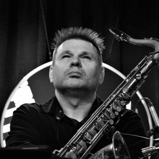 Juodai baltas portretas, kuriame džiazo muzikantas su saksofonu žiūri aukštyn.