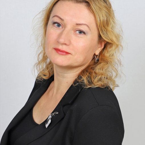 Portretas virš pečių, kuriame moteris su ilgais šviesiais banguotais plaukais, mėlynomis akimis ir rausvais lūpų dažais, dėvi juodą švarką su segėta sage ir juodus auskarus.