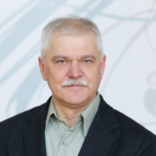 Vyresnio amžiaus vyras su baltais ūsų plaukais, mūvintis juodą švarką ir žalius marškinius, žiūrintis tiesiai į kamerą.