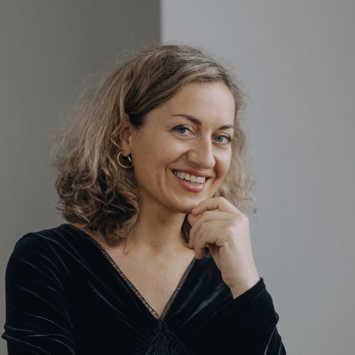 Moteris su garbanotais šviesiais plaukais ir mėlynomis akimis, šypsosi ir laiko ranką prie smakro.