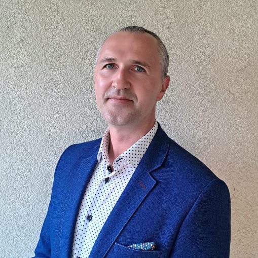 Vyras su mėlynu švarku ir baltiniais su juodomis dėmelėmis stovi prie grublėtos šviesios sienos.