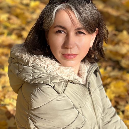 Moteris su šviesiai rudos spalvos striuke su kailine apykakle, stovinti tarp geltonų lapų.