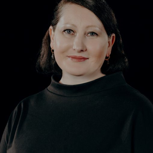 Moteris juodais drabužiais ir tamsiais plaukais, žiūrinti į kamerą.