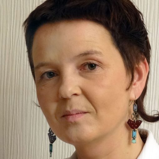 Moters veidas su trumpais tamsiais plaukais ir ilgais auskarais, stovinčios prie raštuotos sienos.