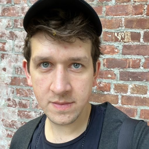 Jaunas vyras su mėlynais akių ir tamsiais plaukais, dėvintis juodą kepurę ir pilką megztinį, stovintis prieš plytų sieną.