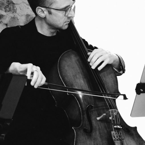 Juodai baltas portretas, kuriame vyras su akiniais griežia violončele.