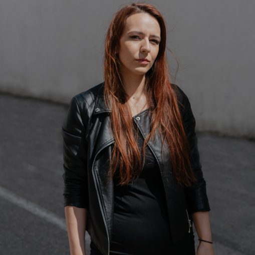 Raudonplaukė moteris vilki juodą odinę striukę ir juodą suknelę, stovinti lauke ant asfalto.