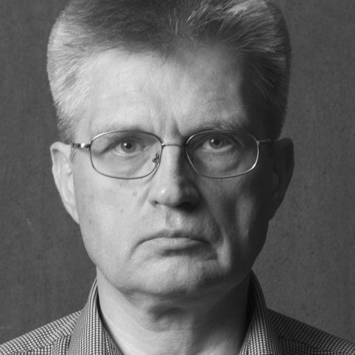 Juodai baltas portretas, kuriame matomas pagyvenęs vyras su akiniais, dėvintis languotus marškinius.