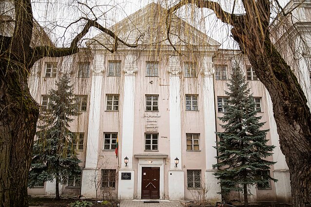 Šviesiai rausvas pastatas su baltais piliastrais, daugybe langų ir mediniu įėjimu, apsuptas šakojusių medžių ir eglių.