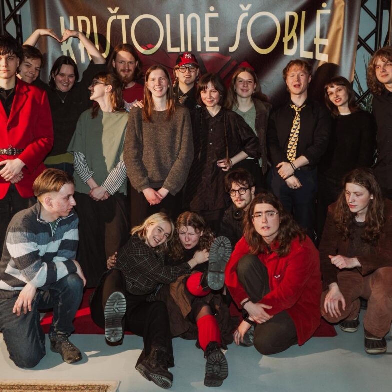 Grupė jaunų žmonių pozuoja nuotraukai prie didelio, šviesaus fono su užrašu "KRITOLINE JOBLĖ".