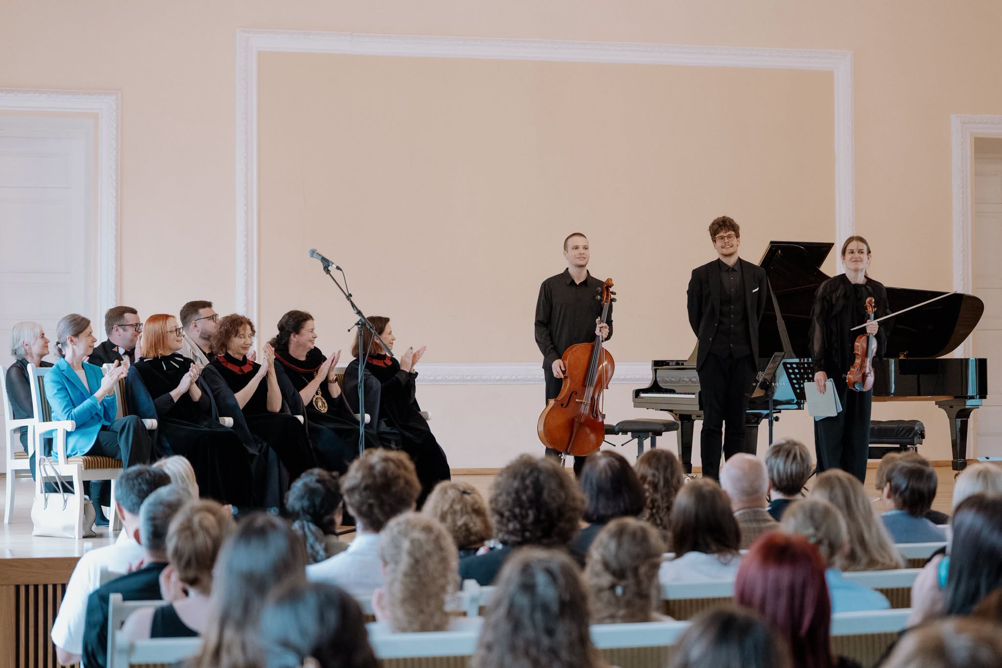 Klasikinės muzikos atlikėjai su instrumentais stovi priešais fortepijoną ir publiką salėje.