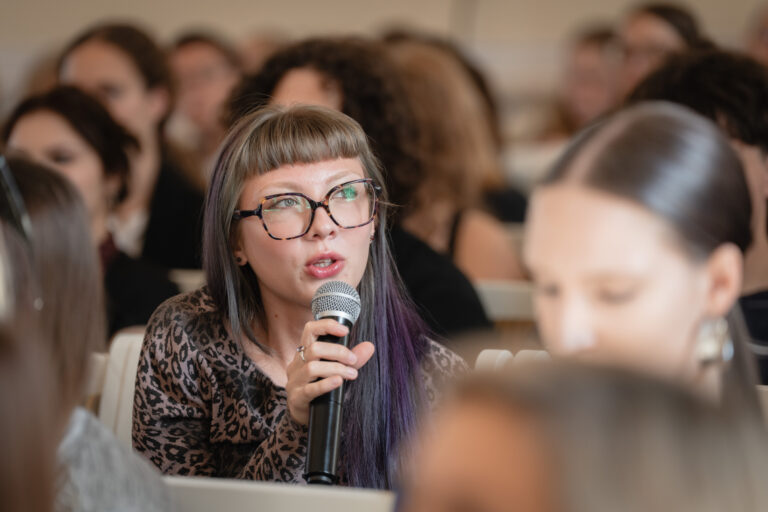 Moteris su akiniais ir purpuriniais plaukų galiukais kalba į mikrofoną, sėdėdama auditorijoje.