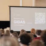 Juodas pianinas ir ekranas su studento gido informacija ir QR kodu, priešais žiūrovų grupę.