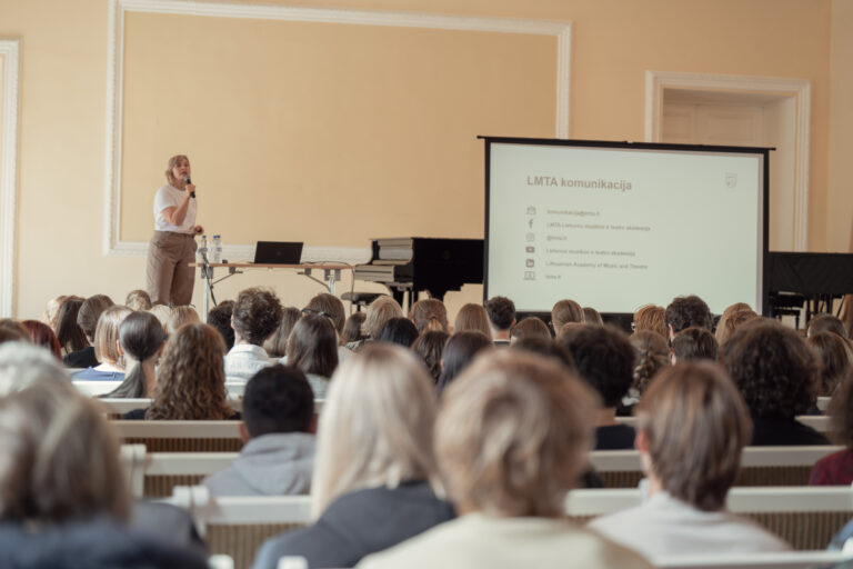 Moteris kalba į mikrofoną prieš auditoriją, kurioje sėdi žmonės, o ekrane matomi LMTA komunikacijos kontaktai.
