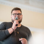 Vyras su akiniais ir barzda kalba į mikrofoną, dėvi pilką megztinį ir pakabuką ant kaklo.