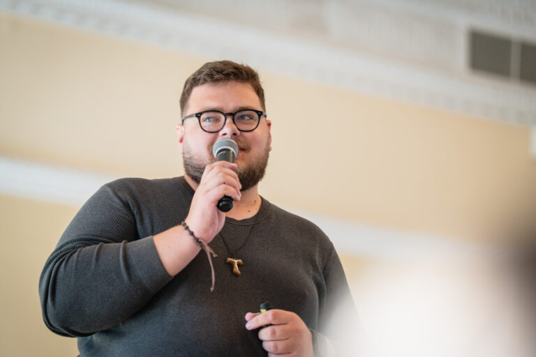 Vyras su akiniais ir barzda kalba į mikrofoną, dėvi pilką megztinį ir pakabuką ant kaklo.