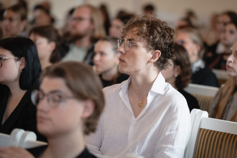 Jaunas vyras su garbanotais rudais plaukais ir akiniais sėdi salėje, dėvi baltus marškinius ir auksinį medalioną.