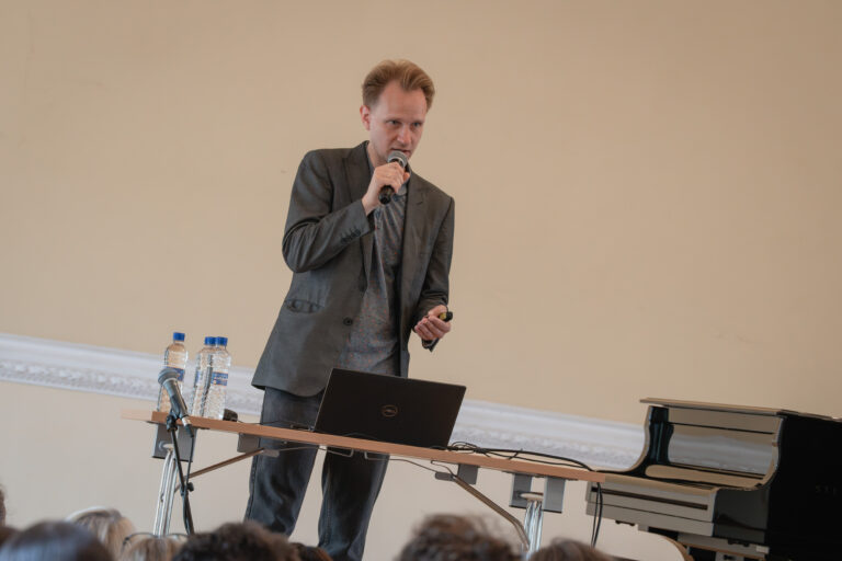 Vyras su akiniais, dėvintis šviesiai pilką švarką ir spalvotus marškinius, kalba į mikrofoną, stovėdamas prie stalo su nešiojamuoju kompiuteriu ir vandens buteliukais, šalia stovi juodas fortepijonas.
