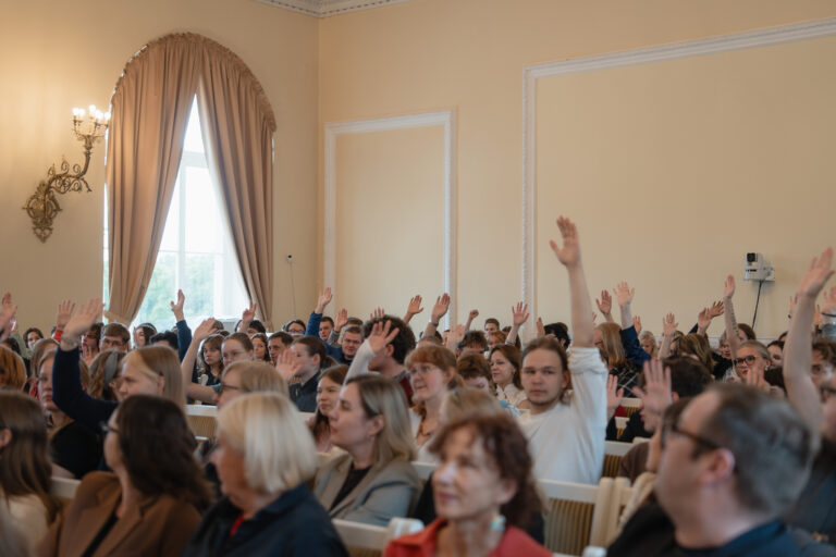 Daug žmonių sėdi auditorijoje ir kelia rankas į viršų.