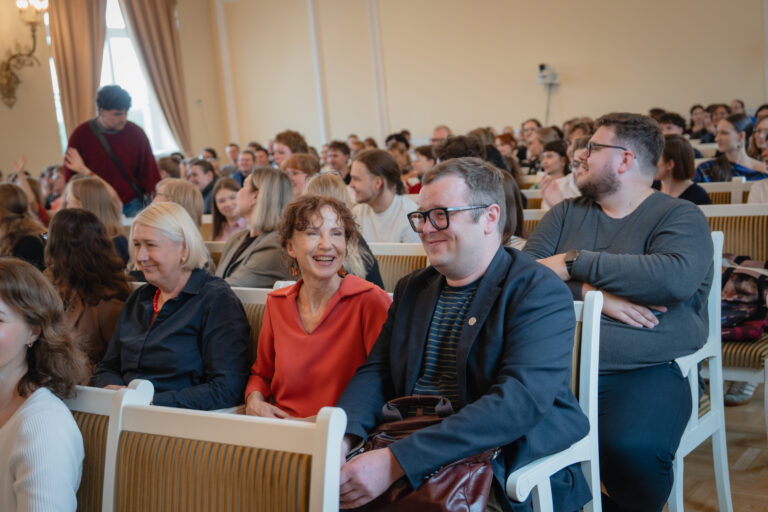 Auditorija su susirinkusiais žmonėmis, dauguma jų sėdi eilėse, klausydamiesi pranešimo, kai kurie iš jų šypsosi ar bendrauja tarpusavyje.
