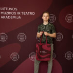Jaunas vyras su pilkšvai žaliu marškinėliais ir džinsais, stovintis prieš tamsiai raudoną foną su LMTA logotipais ir užrašu „LIETUVOS MUZIKOS IR TEATRO AKADEMIJA“, laiko rankose raudoną didelę rankinę su baltomis banguotomis linijomis.