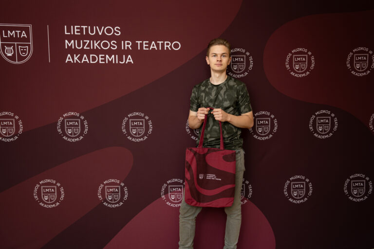 Jaunas vyras su pilkšvai žaliu marškinėliais ir džinsais, stovintis prieš tamsiai raudoną foną su LMTA logotipais ir užrašu „LIETUVOS MUZIKOS IR TEATRO AKADEMIJA“, laiko rankose raudoną didelę rankinę su baltomis banguotomis linijomis.