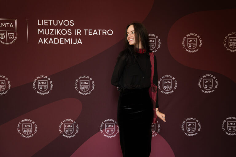 Moteris juoda suknele ir skara šypsosi ant „Lietuvos muzikos ir teatro akademijos“ fono.
