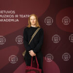 Vyras su ilgais raudonais plaukais ir prakirptais džinsais stovi ant tamsiai raudono fono su baltu užrašu "LIETUVOS MUZIKOS IR TEATRO AKADEMIJA" ir logotipais. Jis dėvi juodus marškinius ir per petį persimetęs rankinę, rankoje laiko raudonų juostų maišelį.