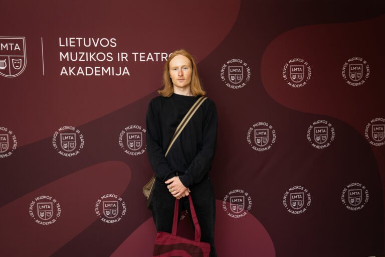 Vyras su ilgais raudonais plaukais ir prakirptais džinsais stovi ant tamsiai raudono fono su baltu užrašu "LIETUVOS MUZIKOS IR TEATRO AKADEMIJA" ir logotipais. Jis dėvi juodus marškinius ir per petį persimetęs rankinę, rankoje laiko raudonų juostų maišelį.