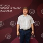 Vyras baltais marškiniais ir mėlynomis kelnėmis stovi prieš tamsiai raudoną foną su baltu užrašu „LIETUVOS MUZIKOS IR TEATRO AKADEMIJA“ ir pakartotinais logotipais.