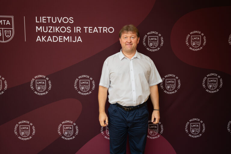 Vyras baltais marškiniais ir mėlynomis kelnėmis stovi prieš tamsiai raudoną foną su baltu užrašu „LIETUVOS MUZIKOS IR TEATRO AKADEMIJA“ ir pakartotinais logotipais.