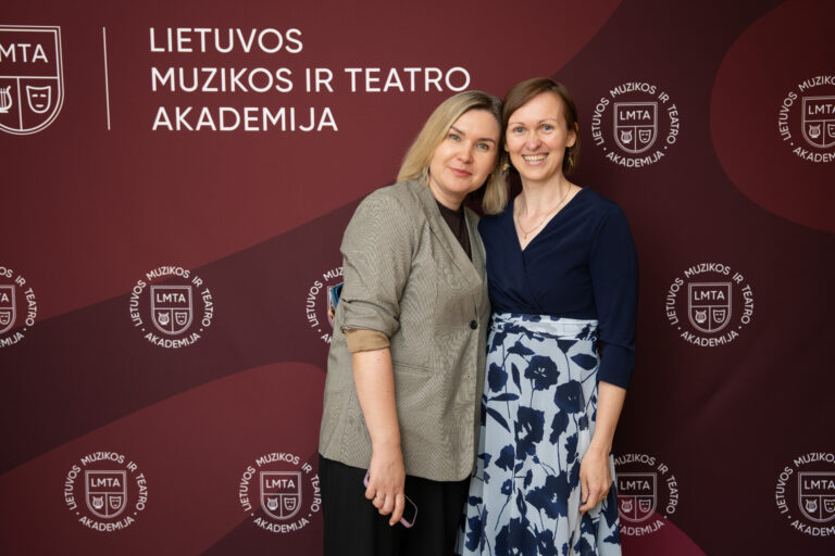Du moterys, viena su languotu švarku ir tamsiu džemperiu, kita su tamsiai mėlyna suknele su gėlėmis, pozuoja ant tamsiai raudono fono su baltu užrašu "LIETUVOS MUZIKOS IR TEATRO AKADEMIJA" ir LMTA logotipais.