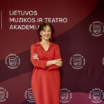 Moteris raudonais drabužiais stovi pasigraibiusi rankas priešais foną su LMTA logotipu ir užrašu "LIETUVOS MUZIKOS IR TEATRO AKADEMIJOS".