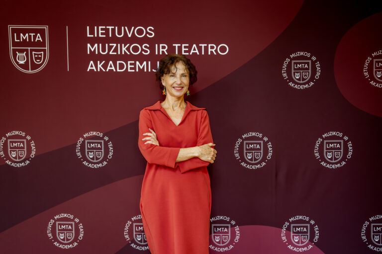 Moteris raudonais drabužiais stovi pasigraibiusi rankas priešais foną su LMTA logotipu ir užrašu "LIETUVOS MUZIKOS IR TEATRO AKADEMIJOS".
