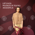 Jauna moteris, vilkinti megztinį ir kelnes, laiko didelį raudoną krepšį su logotipo ir stovi prieš foną su LMTA logotipu ir užrašu "LIETUVOS MUZIKOS IR TEATRO AKADEMIJA".