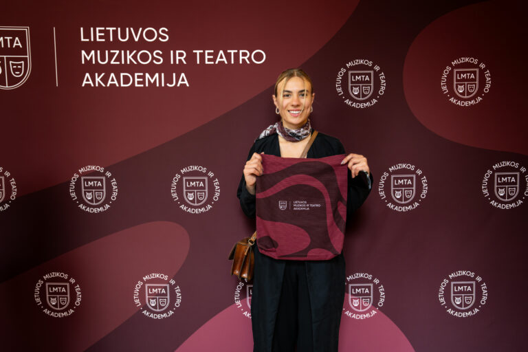 Moteris laiko raudoną rankinę su akademijos logotipu ant tamsiai raudonų sienų fono su logotipais ir kraštovaizdžiu.