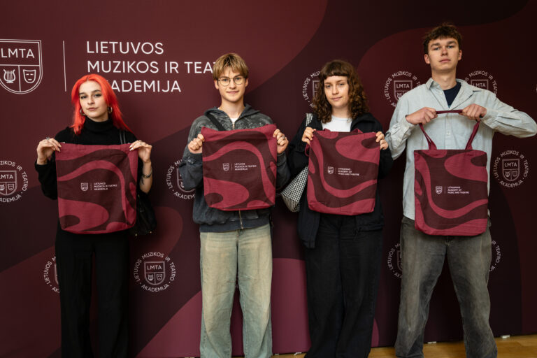 Keturi jauni žmonės stovi prie institucijos, kurios pavadinimas LMTA, logotipais ir užrašais išmargintos sienos, laikydami vienodus, bordinės spalvos su tamsesniais raštais ir LMTA logotipu maišelius.