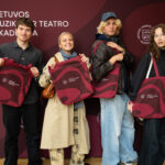 Keturi jauni žmonės, du vyrai ir dvi moterys, stovi prieš raudoną foną su baltu užrašu "LIETUVOS MUZIKOS IR TEATRO AKADEMIJA" ir laiko rankose didelius raudonus krepšius.