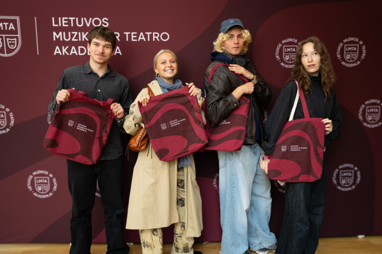 Keturi jauni žmonės, du vyrai ir dvi moterys, stovi prieš raudoną foną su baltu užrašu "LIETUVOS MUZIKOS IR TEATRO AKADEMIJA" ir laiko rankose didelius raudonus krepšius.