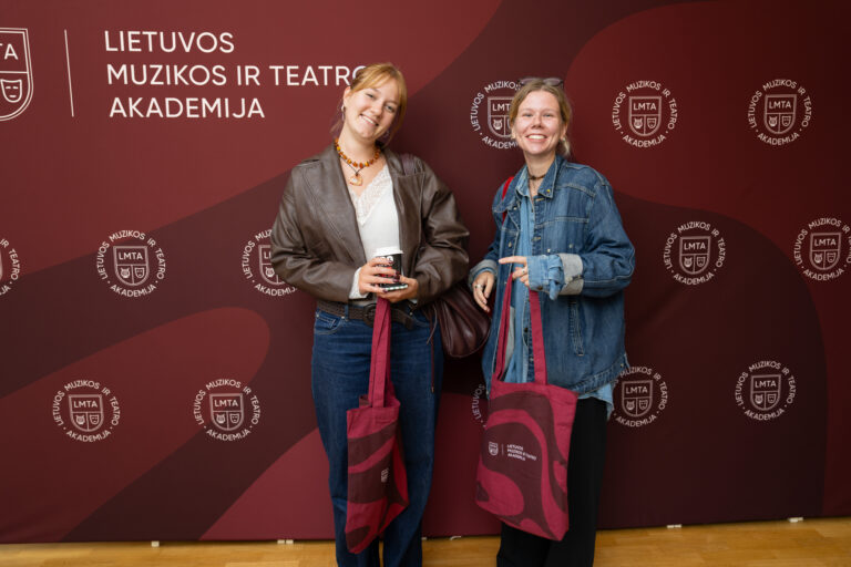 Du jauni moterys stovi prieš rudą foną su logotipais, viena laiko puodelį kavos, o kita rodo ranka.