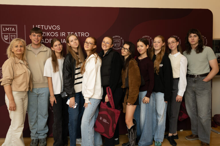 A group of ten young people and one older woman stand in front of a maroon banner with white text and logos.