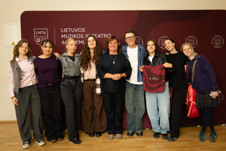 Grupė žmonių, tarp jų moteris ir kelios merginos bei vienas vaikinas, stovi prieš tamsiai raudoną foną su LMTA logotipais ir užrašu „LIETUVOS MUZIKOS IR TEATRO AKADEMIJA“. Nuotrauka daryta horizontaliai, žmonės šypsosi.