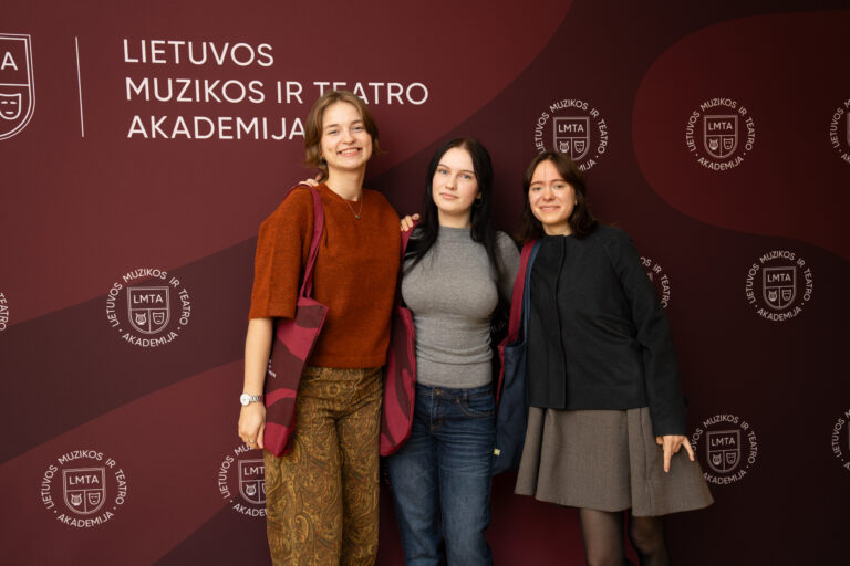 Trys jaunos moterys stovi prieš rudą foną su baltu užrašu „Lietuvos muzikos ir teatro akademija“ ir logotipais.